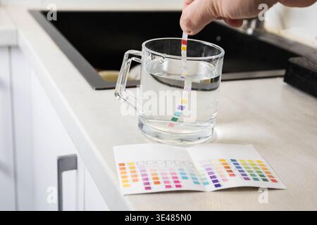Nahaufnahme der Hand, die den chemischen Teststreifen hält und ihn in einen mit Wasser gefüllten Klarglasbecher eintaucht, der sich auf der Küchenarbeitsplatte befindet. Farbverweis Stockfoto