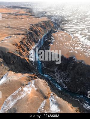 Aus der Vogelperspektive auf einen tiefen Canyon mit einem sich windenden türkisfarbenen Fluss, der zwischen steilen Felsklippen und schneebedeckten Hochplateaus in Kasachstan fließt. Stockfoto