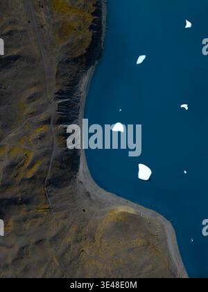 Aus der Vogelperspektive schwimmende weiße Eisberge im tiefblauen Gletscherwasser entlang einer dunklen vulkanischen Küste Islands. Stockfoto