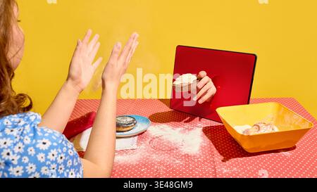 Frau klatscht in die Hände, während auf dem Tablettenschirm auf dem mit Mehl bedeckten Tisch Torte erscheint. Stockfoto
