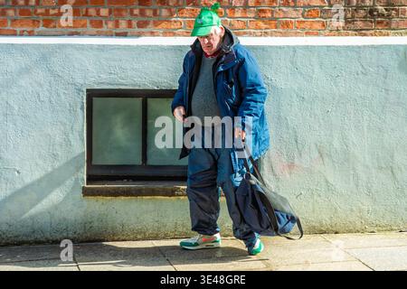Ein älterer einheimischer Mann in alltäglicher Kleidung mit einer Shamrock-Mütze spaziert am St. Patrick’s Day durch das Stadtzentrum von Dublin und feiert auf subtile Weise das Festival am 17. März. Stockfoto