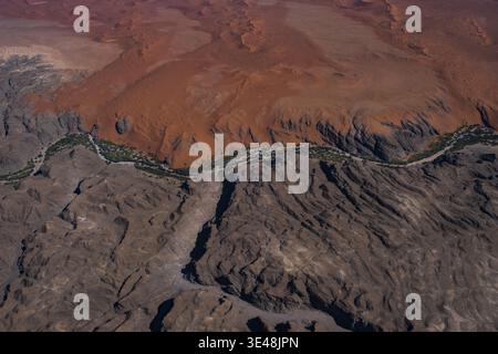 Aus der Vogelperspektive sehen Sie den beeindruckenden Kontrast zwischen roten Sanddünen und dunklen felsigen Bergen mit einer dünnen Linie grüner Vegetation in Walvis Bay, Erongo Region Stockfoto