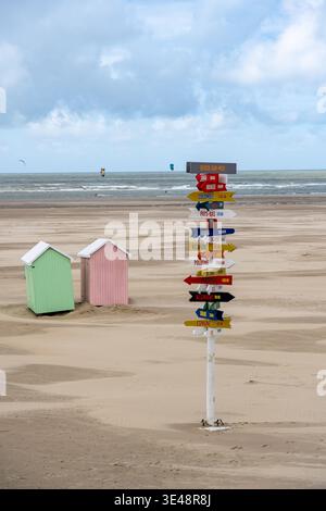 Blick auf farbenfrohe Richtungsschilder, die in alle Richtungen zeigen, hoch am Sandstrand mit pastellfarbenen Strandhütten, unter bewölktem Himmel, Berck-Plage, Stockfoto