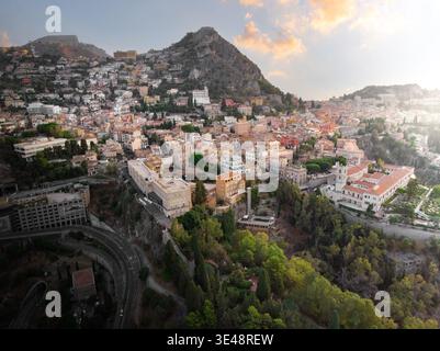 Blick aus der Vogelperspektive auf die historische Stadt am Hügel mit traditionellen Dächern, üppigem Grün und einer gewundenen Straße unter dem goldenen Sonnenuntergang Taormina, Sizilien, Ita Stockfoto