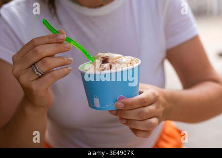 Unerkennbare junge Frau, die ein lässiges weißes T-Shirt trägt, das eine Papierschale mit einem Eislöffel hält. Sommer-Lifestyle-Porträt mit Fokus auf weibliche ha Stockfoto