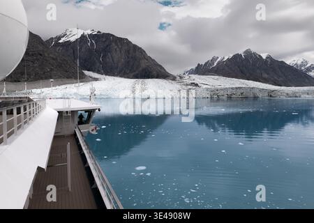 Blick auf einen massiven Gletscher, der in das blaue Meer mit schneebedeckten Bergen unter einem bewölkten Himmel fließt, von einem Kreuzfahrtschiff Grönland aus gesehen. Stockfoto