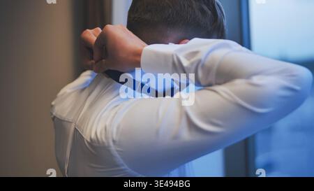 Der Bräutigam macht sich bereit für die Hochzeit und passt seine blaue Fliege vor einem Fenster an. Rückansicht. Stockfoto