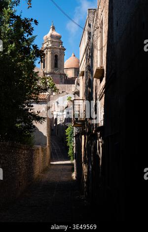 Blick auf eine enge Kopfsteinpflasterallee, die zu einem historischen Kirchturm und einer Kuppel unter einem klaren blauen Himmel in Segesta, Sizilien, Italien führt. Stockfoto