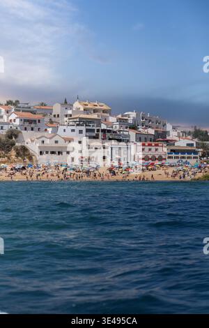 Blick auf einen überfüllten Strand mit farbenfrohen Sonnenschirmen und weißen Häusern auf dem Hügel mit Blick auf das blaue Meer in Portimao, Faro District, Portugal. Stockfoto