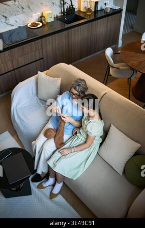 Vertikale Aufnahme einer älteren Frau, die Mädchen beibringt, die zu Hause auf dem Sofa strickt, Familienhobbys und praktische Fähigkeiten teilt. Geeignet für Elternschaft, intergenerationelle Bindung und Lifestyle-Kampagnen Stockfoto