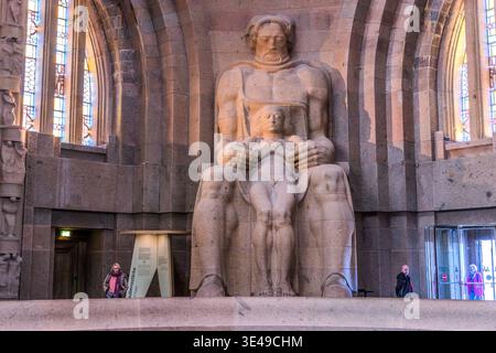 Krypta des Monuments für die Völkerschlacht, allgemeine Ansicht: Eine monumentale Figur mit einem Kind vor den Buntglasfenstern; Besucher im Vordergrund vermitteln ein Gefühl der Skala. Leipzig, Sachsen, Deutschland Stockfoto