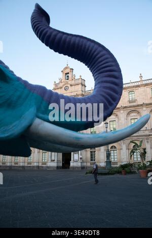 Blick auf einen farbenfrohen Elefanten-Skulpturenkasten und Stoßzähne vor dem historischen Palazzo degli Elefanti mit einem Mann, der über den Platz Catania, Si, spaziert Stockfoto