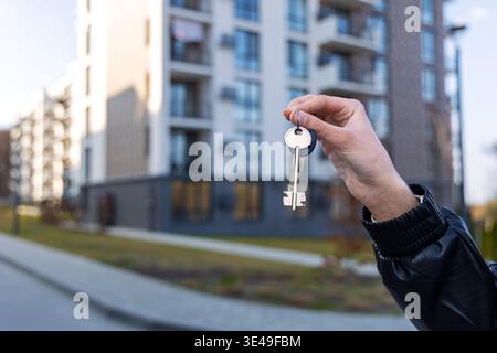 Hand Hält Die Wohnungsschlüssel Vor Dem Neubau. Nahaufnahme einer Hand mit Hausschlüsseln vor einem modernen Wohnhaus, das ein neues Zuhause symbolisiert Stockfoto