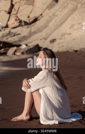 Junge Frau, die auf Strandsand sitzt, Profilansicht, entspannt und reflektiert mit geschlossenen Augen Stockfoto