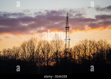 Radioantenne und Sendemast, die sich in der Abenddämmerung über einer Waldlinie in der lettischen Landschaft erheben, mit orangefarbenen Wolken und Industrietechnik Stockfoto