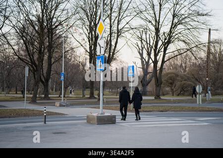 Rückansicht eines Paares in dunklen Mänteln, das auf einem Quersteg in einem Rigaer Park spaziert, mit Fußgängerschildern und städtischem Umfeld für das europäische Stadtleben Stockfoto