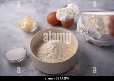 Nahaufnahme der Backzubereitung auf einer neutral strukturierten Arbeitsplatte: Keramikschale mit Mehl, Löffel Schaufelmehl, braune Eier, Buttertupfer und s Stockfoto
