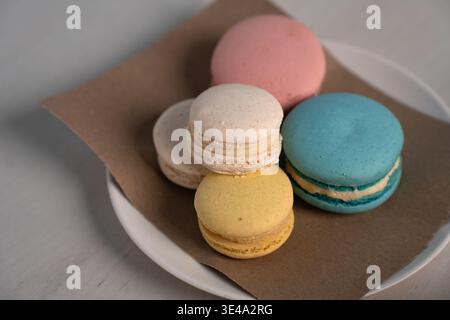 Nahaufnahme von fünf pastellfarbenen Macarons, die auf braunem Pergament über einer weißen Platte und einer hellen Tischplatte angeordnet sind und zarte Schalen und cremige Füllungen hervorheben Stockfoto