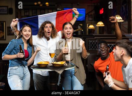 Fröhliche, multirassische Fußballfans schwenken in der Sportbar mit Bier und Chips die Flagge Frankreichs Stockfoto