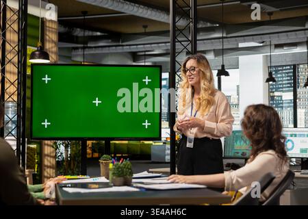 Ein Team von Geschäftsfrauen erstellt neben dem Chroma Key Display eine Strategie mit einem großen TV-Monitor, der offen ist und Planungsdokumente auf dem Tisch liegen. Mitarbeiter zeigen die Effizienz von Geschäftsabläufen. Stockfoto