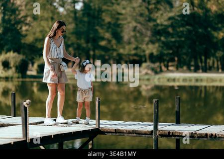 Mutter und Tochter halten Hände, während sie einen gemütlichen Spaziergang auf einem hölzernen Dock in einem malerischen Seeufer genießen Stockfoto