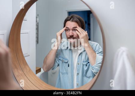 Selbstbewusster Mann, der auf die Reflexion im Badezimmerspiegel blickt und die Gesichtshaut mit einem zufriedenen Lächeln berührt. Stockfoto