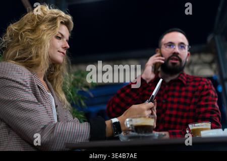 Zwangloses Treffen mit einer blonden Frau und einem bärtigen Mann in gemütlicher Café-Umgebung, Besprechung der Arbeit und Kaffeegenuss unter warmer Umgebungsbeleuchtung Stockfoto