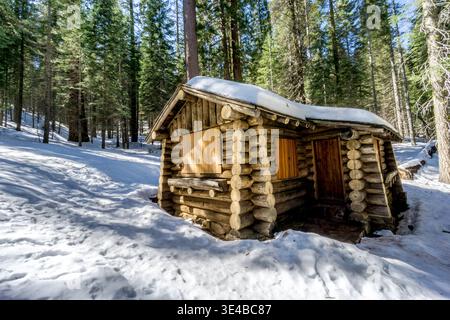Eine kleine Blockhütte ist mit Schnee bedeckt. Die Hütte ist von Bäumen umgeben und liegt in einem verschneiten Wald Stockfoto