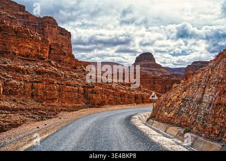Die gewundene Asphaltstraße, die durch die zerklüfteten rötlichen Felsformationen und die hohen Klippen der trockenen Wüstenlandschaft unter einem bewölkten Himmel navigiert, schlägt vor Stockfoto