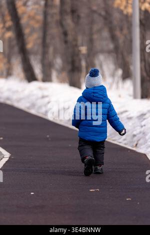 Ein kleines Kind in einer hellblauen Winterjacke spaziert über einen verschneiten Pfad in einem Park Stockfoto