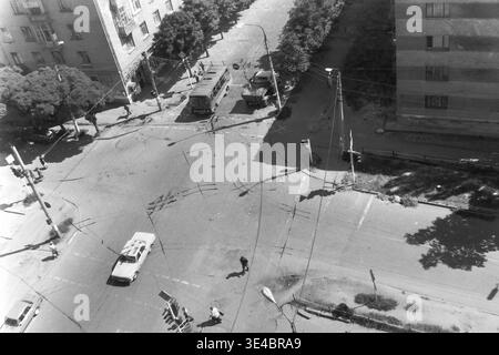 Slawjansk, 1998, Kreuzung der Straßen Oktjabrskoj Revolutsii (heute Wasyliwska) und Yunykh Kommunarow (heute Bankiwska). Archivieren Sie ein Schwarzweißfoto, das von oben nach unten eine geschäftige Straßenkreuzung von einem neunstöckigen Gebäude aus aufnimmt. Die detailreiche Szene zeigt einen vorbeifahrenden Bus, Oldtimer, Fußgänger, die die Asphaltstraße überqueren, und Obus-Drähte. Dieses dynamische architektonische Bild dokumentiert den alltäglichen Stadtverkehr, die lokale Verkehrsinfrastruktur und den friedlichen Donbass perfekt. Stockfoto