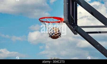 Basketball schießt einen Korb, schwimmt durch das Netz vor einem bewölkten blauen Himmel. Stockfoto
