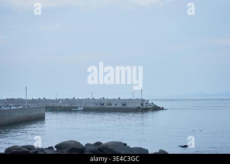 Ein ruhiger kleiner Hafen mit einem schwimmenden gelben Boot, einem roten Auto und Menschen, die einen entspannten Tag mit Angeln unter einem ruhigen, weichen, bedeckten Himmel genießen. Stockfoto