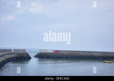 Ein ruhiger kleiner Hafen mit einem schwimmenden gelben Boot, einem roten Auto und Menschen, die einen entspannten Tag mit Angeln unter einem ruhigen, weichen, bedeckten Himmel genießen. Stockfoto