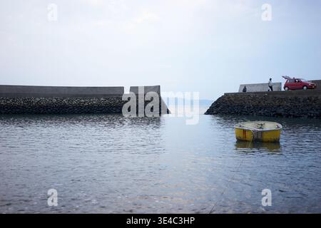 Ein ruhiger kleiner Hafen mit einem schwimmenden gelben Boot, einem roten Auto und Menschen, die einen entspannten Tag mit Angeln unter einem ruhigen, weichen, bedeckten Himmel genießen. Stockfoto