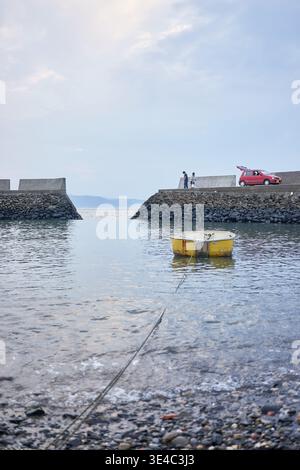 Ein ruhiger kleiner Hafen mit einem schwimmenden gelben Boot, einem roten Auto und Menschen, die einen entspannten Tag mit Angeln unter einem ruhigen, weichen, bedeckten Himmel genießen. Stockfoto