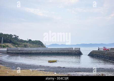 Ein ruhiger kleiner Hafen mit einem schwimmenden gelben Boot, einem roten Auto und Menschen, die einen entspannten Tag mit Angeln unter einem ruhigen, weichen, bedeckten Himmel genießen. Stockfoto