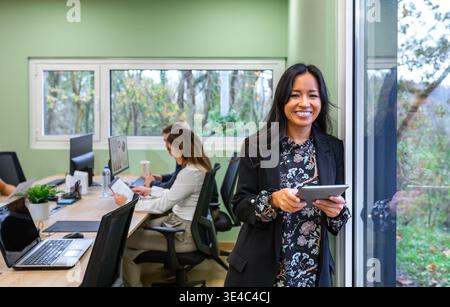 Mittlere Aufnahme einer lächelnden hispanischen Geschäftsfrau, die ein digitales Tablet vor einem Bürofenster hält Stockfoto