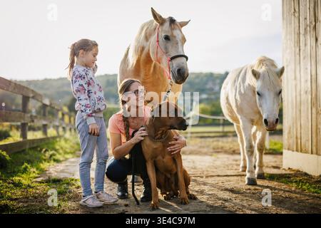 Porträt der Frau und ihrer Tochter mit Hund und Pferden auf der Familienfarm bei Sonnenuntergang. Ländlicher Lebensstil mit Haustieren und Tieren Stockfoto