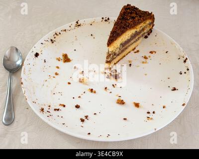 Das letzte Stück mehrschichtiger Gluten- und laktosefreier Biskuitkuchen, der auf einer weißen Platte mit Krümeln neben einem Teelöffel verbleibt. Stockfoto