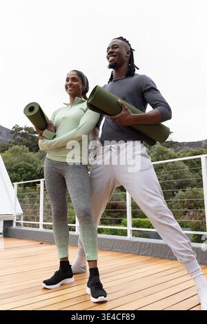 Verschiedene Freunde stehen auf einer erhöhten Holzterrasse mit gerollten grünen Yogamatten und tragen Aktivkleidung. Outdoor, Terrasse, Fitness, Duo, Natur, bewölkt, lässig || Model veröffentlicht Stockfoto
