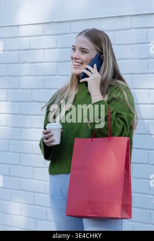 Frau in grünem Pullover, die auf blauem Smartphone spricht, rote Tasche hält, Tasse gegen weiße Ziegelwand Stockfoto