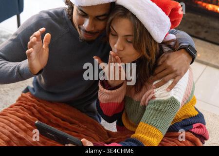 Ein Paar teilt sich eine orange gesteppte Decke am Kamin, hält eine schwarze Tafel und trägt Santa-Hüte Stockfoto