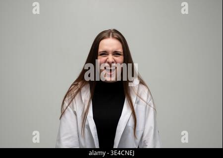 Junge frustrierte Frau, die mit offenem Mund schreien und Zorn und Aggression vor grauem Hintergrund ausdrücken Stockfoto