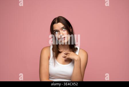 Junge Frau in einem weißen Oberteil, die mit breiten Augen und offenem Mund aufblickt, Überraschung ausdrückt oder über ein Konzept auf rosa Hintergrund nachdenkt Stockfoto