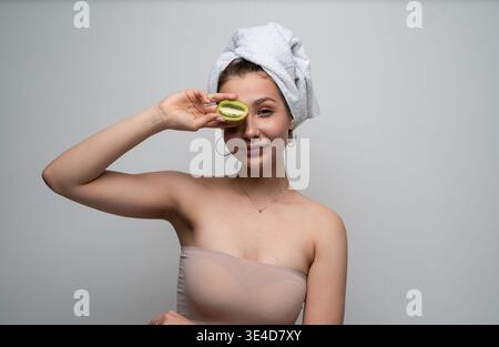 Junge Frau, die natürliche Schönheit und gesunde Hautkonzepte präsentiert und nach dem Duschen eine halbe Kiwi-Frucht über einem Auge hält, bereit für eine Hautpflege-Routine Stockfoto