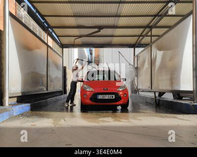 Junge Frau, die auf Zehenspitzen stand, mit Hochdruckwasserpistole, um das rote Ford Ka Kompaktwagendach in der Selbstbedienungsanlage der städtischen Waschanlage zu reinigen, Vorderansicht, Klimaanlage Stockfoto