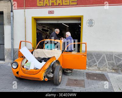 Zwei ältere Männer arbeiten an der Restaurierung eines zerlegten orangen Volkswagen Käfers Typ 1 vor einer Garage, umgeben von Werkzeugen und Teilen in Barcelona. Stockfoto