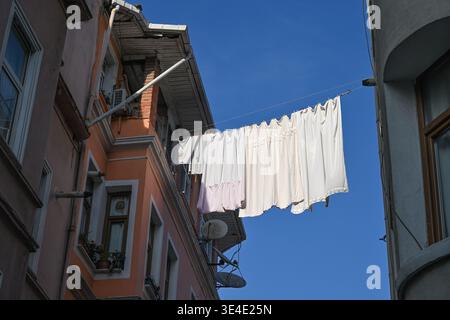 Weiße Bettwäsche wird auf einer Wäscheleine getrocknet, die sich zwischen Häusern vor einem klaren, hellblauen Himmel erstreckt. Istanbul, Türkei Stockfoto
