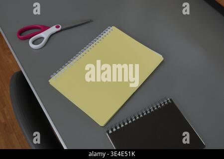 Minimalistische Schreibtischszene mit Werkzeugen und Notebooks. Die Schere liegt neben zwei spiralgebundenen Notizbüchern. Gelbe und schwarze Abdeckungen stehen im Kontrast zur grauen Oberfläche Stockfoto